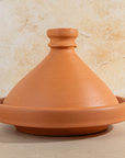 A terracotta clay tagine pot with a matching conical lid, set against a textured beige stone wall background.