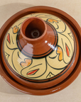 A round ceramic lidded pot or tagine with a terracotta brown glazed body and a cream-colored patterned top featuring black and orange decorative motifs.