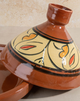 A traditional Moroccan ceramic tagine pot with its matching lid resting beside it. The tagine features a glossy terracotta brown glazed body with a beige glazed interior, decorated with black floral outlines and orange and red accents.
