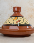 A traditional Moroccan ceramic tagine pot with a glazed terracotta brown lid and base, featuring a cream-colored painted belly with black, yellow, and red abstract patterns, set against a textured beige stone background.