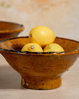 A rustic handmade ceramic bowl with a footed base, filled with three yellow lemons, set against a warm beige background with another similar bowl partially visible in the soft-focus background.