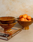 Three stacked rustic brown ceramic bowls with a glossy finish, displayed on a book with oranges in a bowl, against a textured beige wall.