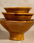 Three nested ceramic bowls with a rustic, glossy ochre-brown glaze and dark speckles, set against a textured beige background.