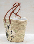 Woven basket with leather handles and embroidered design on a light background