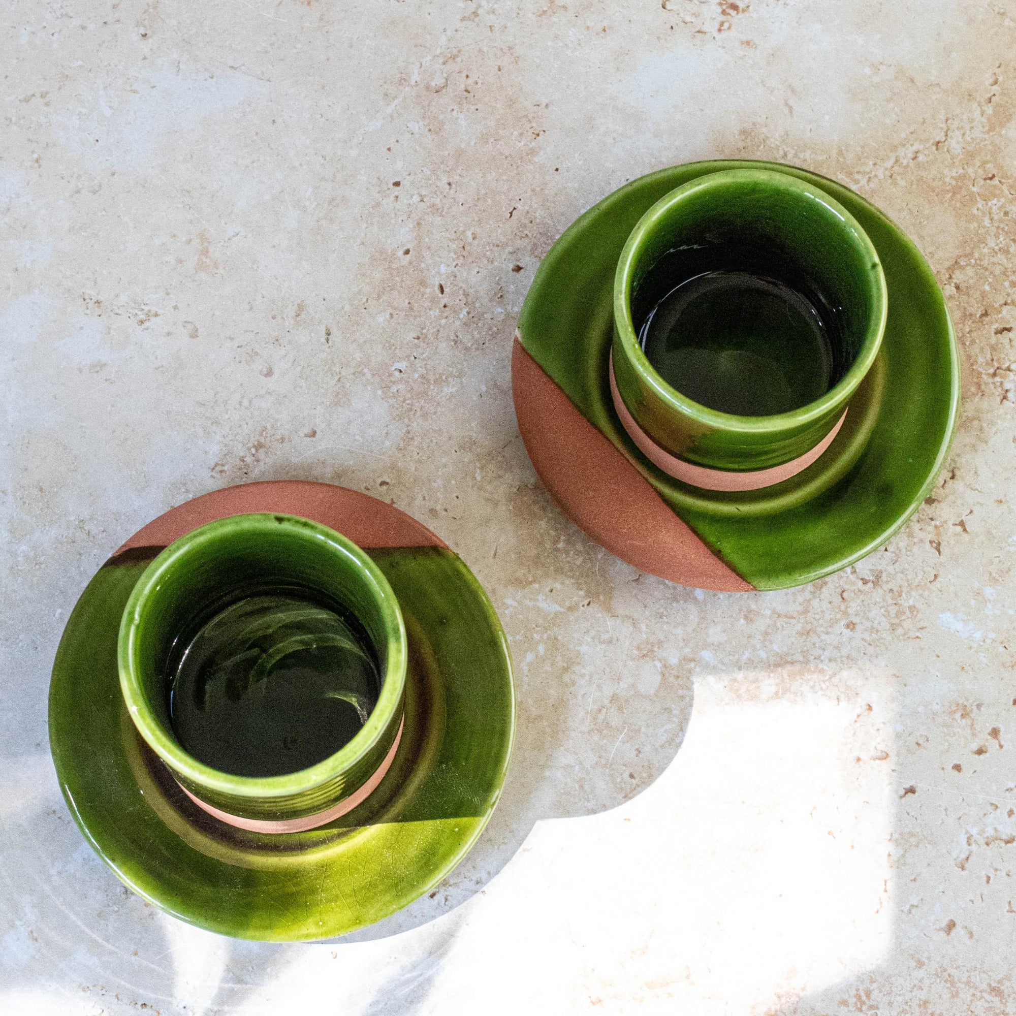 Two green ceramic cups with terracotta bases on a beige surface