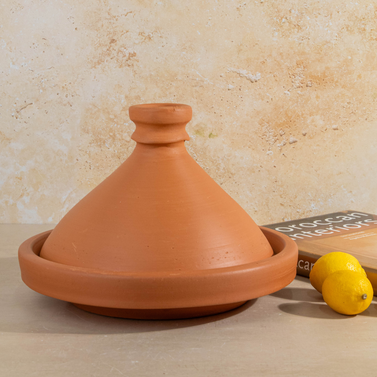 A terracotta clay tagine pot with a matching shallow saucer, shown on a tabletop against a textured beige wall. Two lemons and part of a book are placed beside it as styling props.