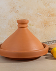 A terracotta clay tagine pot with a matching shallow saucer, shown on a tabletop against a textured beige wall. Two lemons and part of a book are placed beside it as styling props.