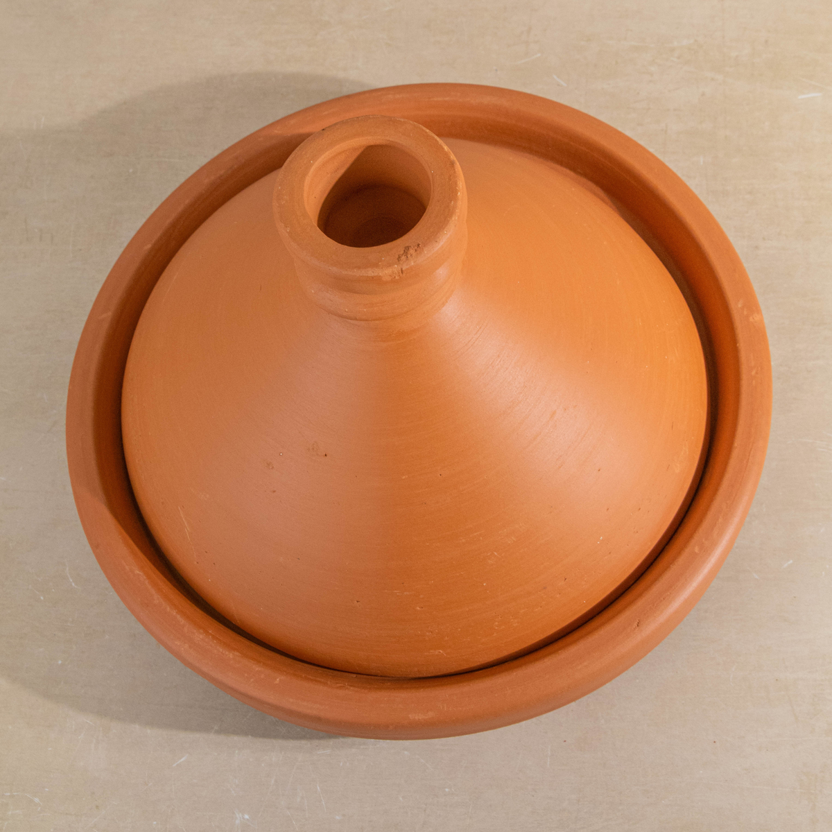 A terracotta clay tagine pot with a matching lid, photographed from directly above on a light beige surface.