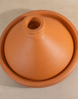 A terracotta clay tagine pot with a matching lid, photographed from directly above on a light beige surface.