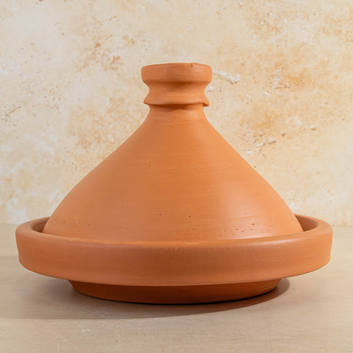 A terracotta clay tagine pot with a matching conical lid, set against a textured beige stone wall background.