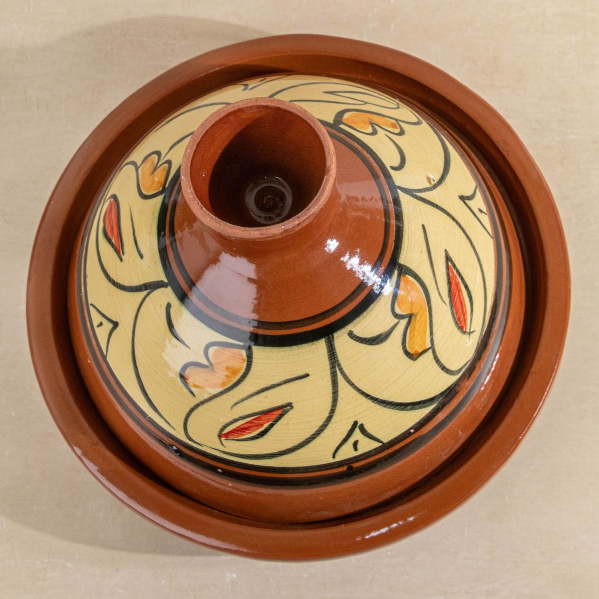 A round ceramic lidded pot or tagine with a terracotta brown glazed body and a cream-colored patterned top featuring black and orange decorative motifs.