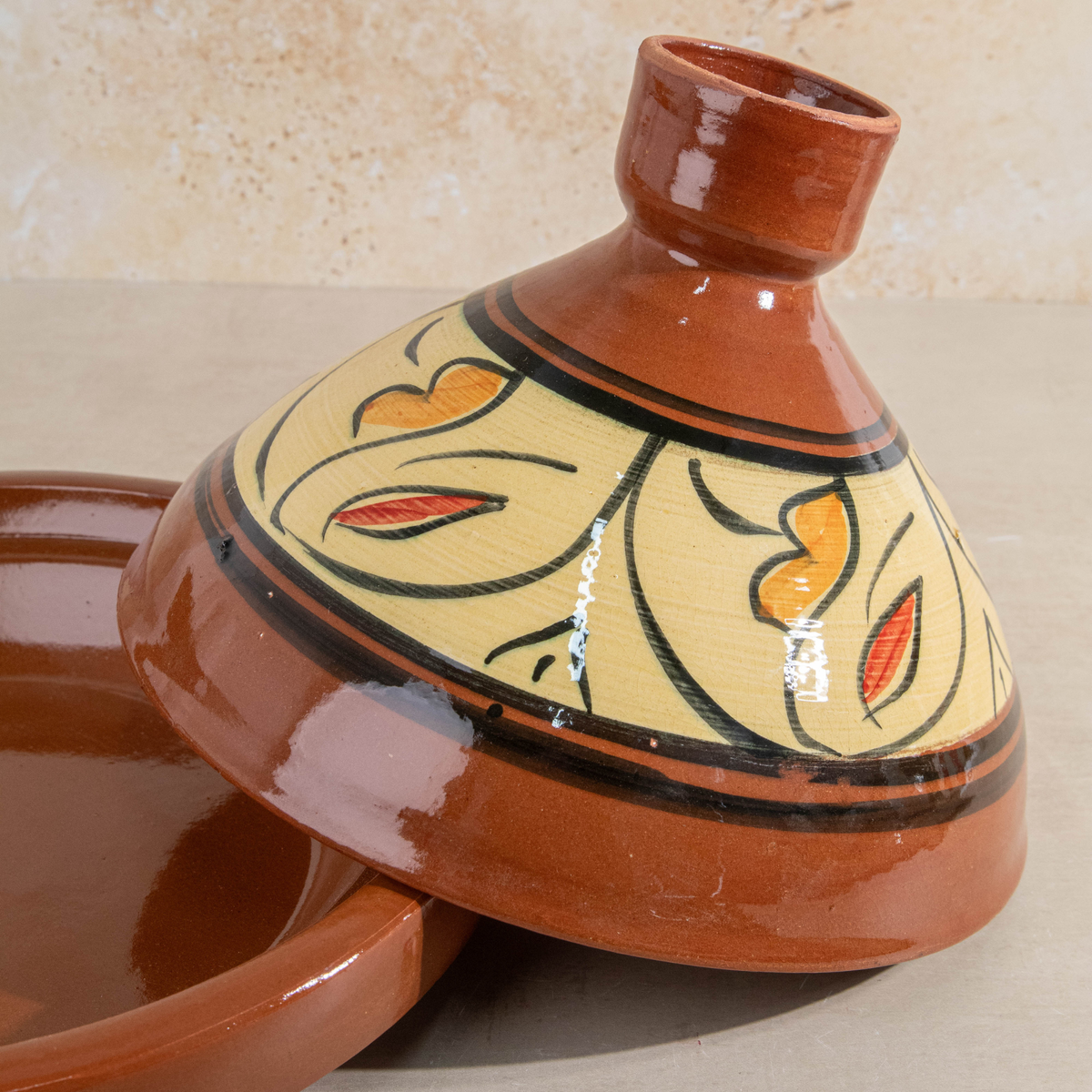 A traditional Moroccan ceramic tagine pot with its matching lid resting beside it. The tagine features a glossy terracotta brown glazed body with a beige glazed interior, decorated with black floral outlines and orange and red accents.