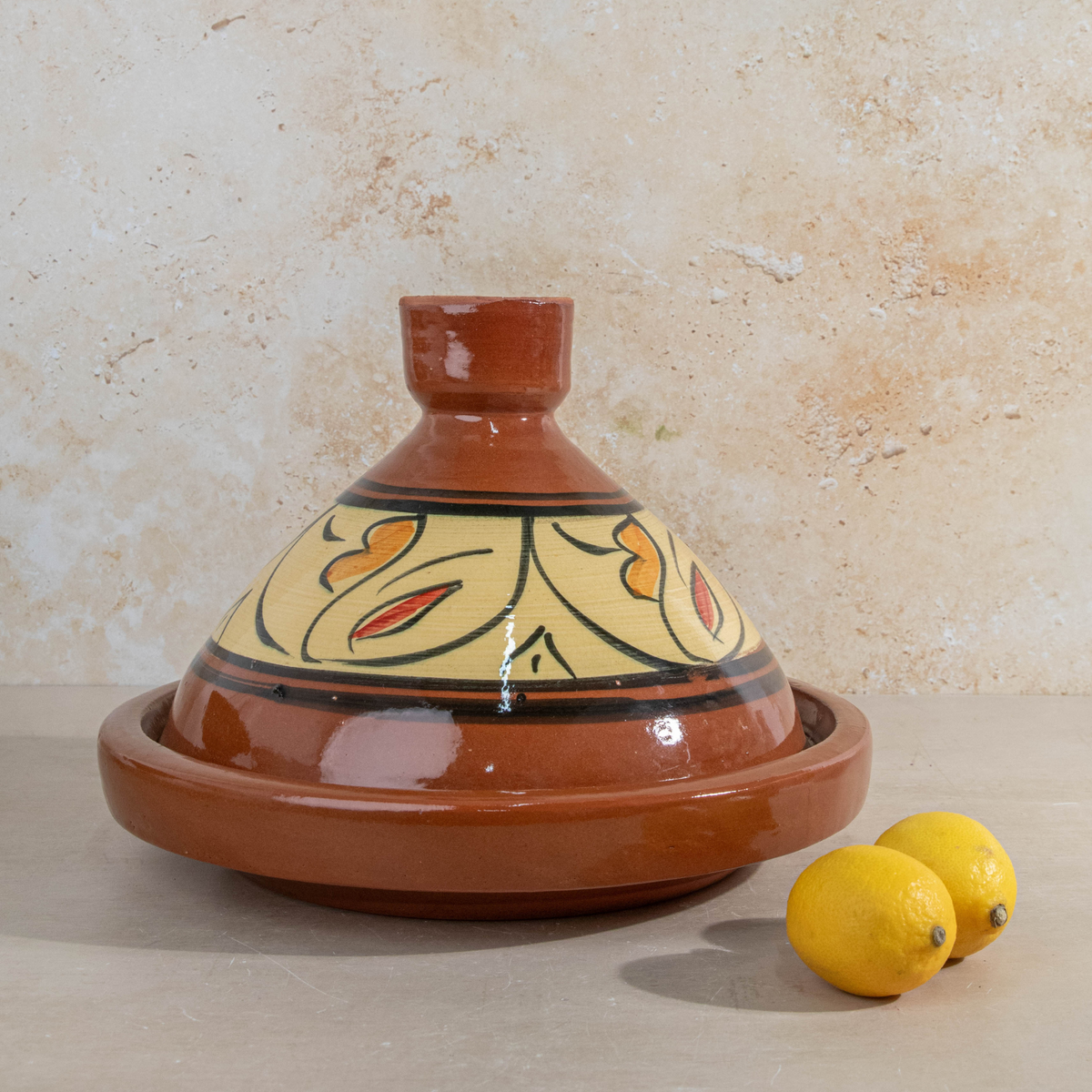 A glazed terracotta tagine pot with a conical lid, decorated with a yellow, orange, and black floral pattern, shown on a beige stone surface with two lemons in the foreground.