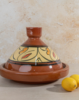 A glazed terracotta tagine pot with a conical lid, decorated with a yellow, orange, and black floral pattern, shown on a beige stone surface with two lemons in the foreground.