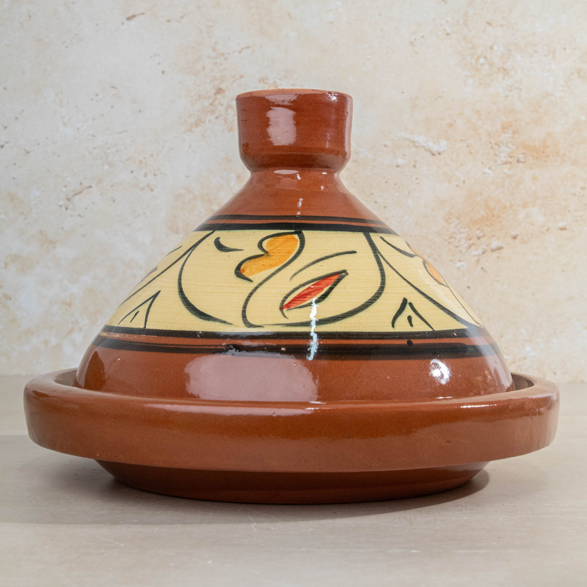 A traditional Moroccan ceramic tagine pot with a glazed terracotta brown lid and base, featuring a cream-colored painted belly with black, yellow, and red abstract patterns, set against a textured beige stone background.