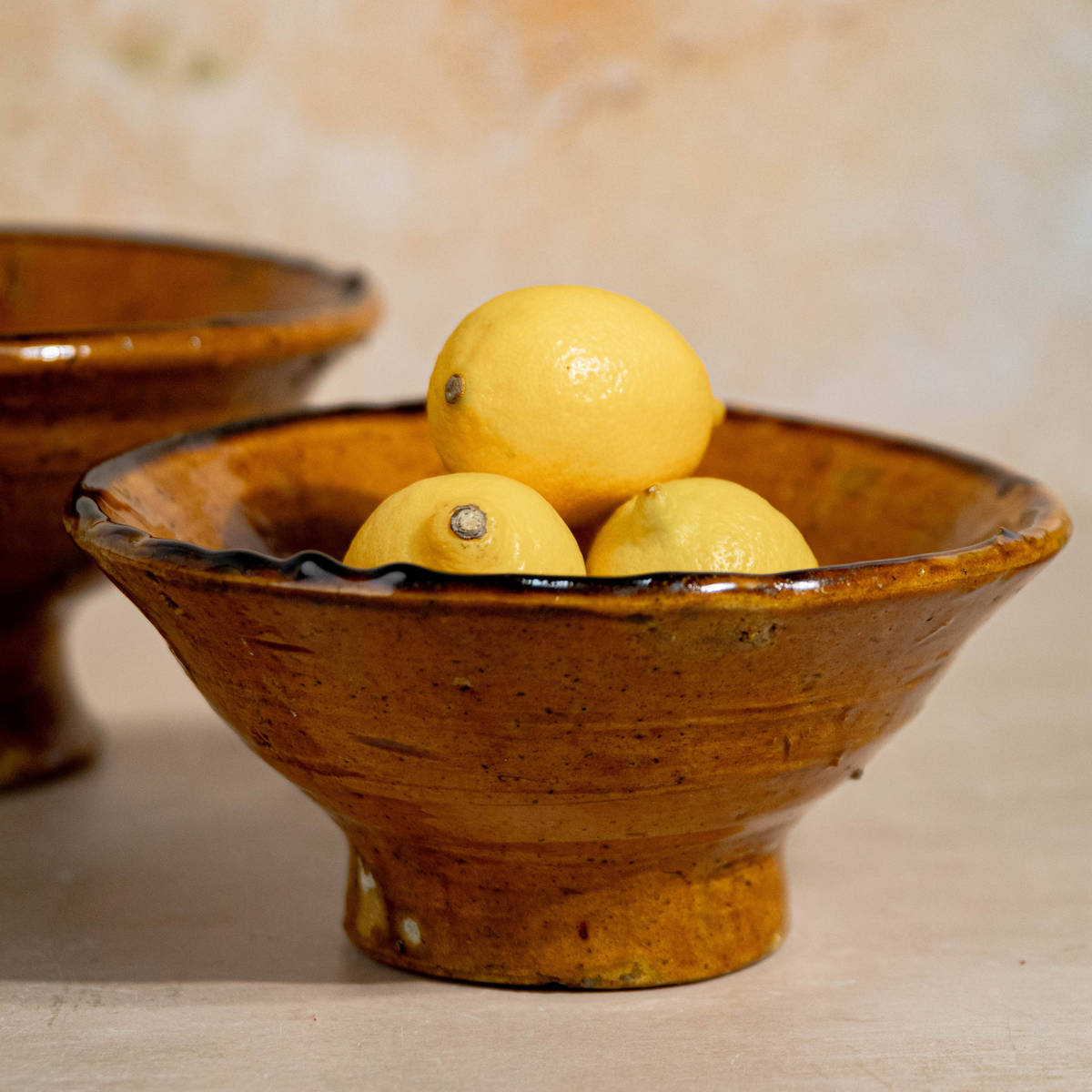 A rustic handmade ceramic bowl with a footed base, filled with three yellow lemons, set against a warm beige background with another similar bowl partially visible in the soft-focus background.