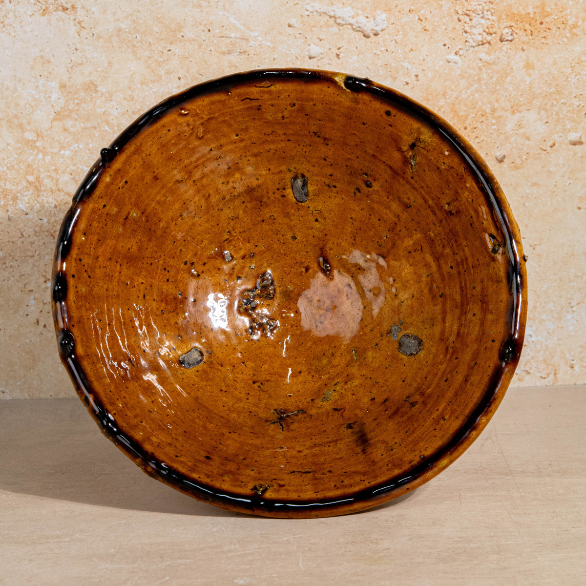 A handmade ceramic bowl with a rustic, hand-painted finish, featuring a warm brown and orange glaze with dark brown speckles and a dark rim, set against a textured beige background.