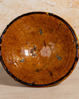 A handmade ceramic bowl with a rustic, hand-painted finish, featuring a warm brown and orange glaze with dark brown speckles and a dark rim, set against a textured beige background.