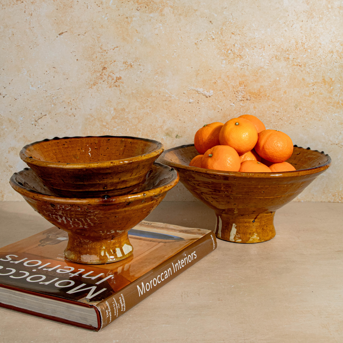 Three stacked rustic brown ceramic bowls with a glossy finish, displayed on a book with oranges in a bowl, against a textured beige wall.