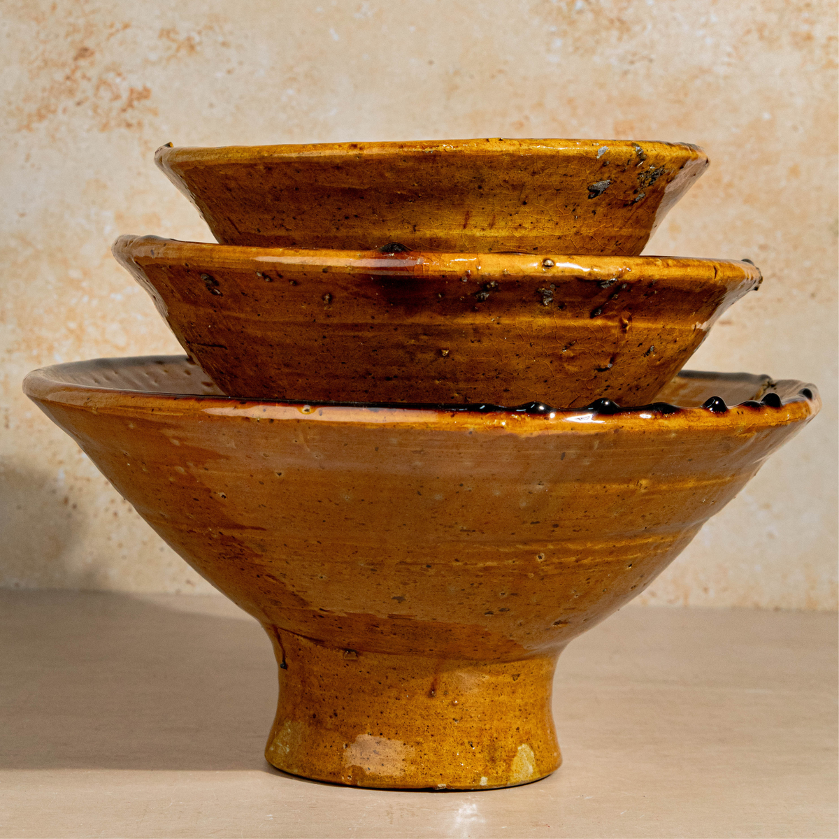 Three nested ceramic bowls with a rustic, glossy ochre-brown glaze and dark speckles, set against a textured beige background.