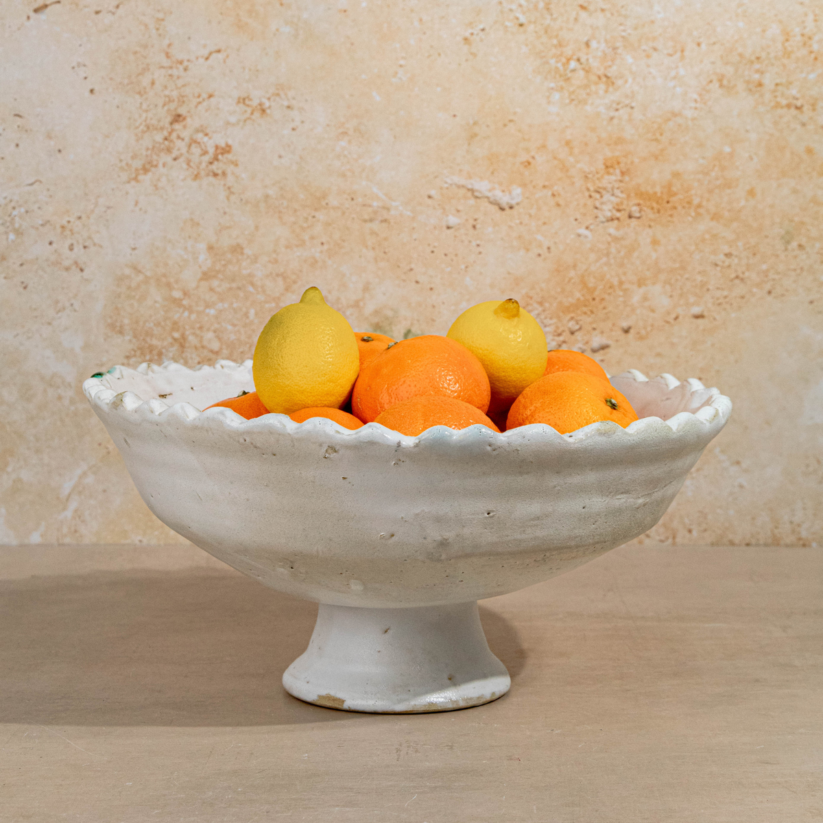 A white ceramic pedestal bowl with a scalloped rim, filled with oranges and lemons, set against a textured beige stone wall and wooden surface.