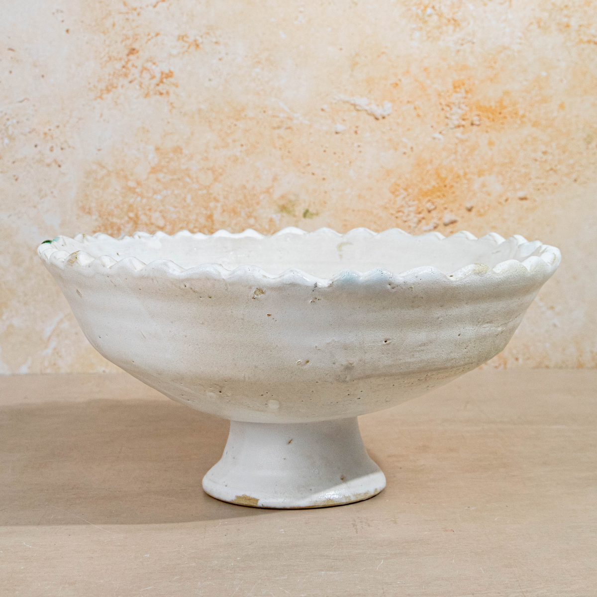 An off-white ceramic pedestal bowl with a scalloped rim, set against a textured beige stone wall and light wood surface.