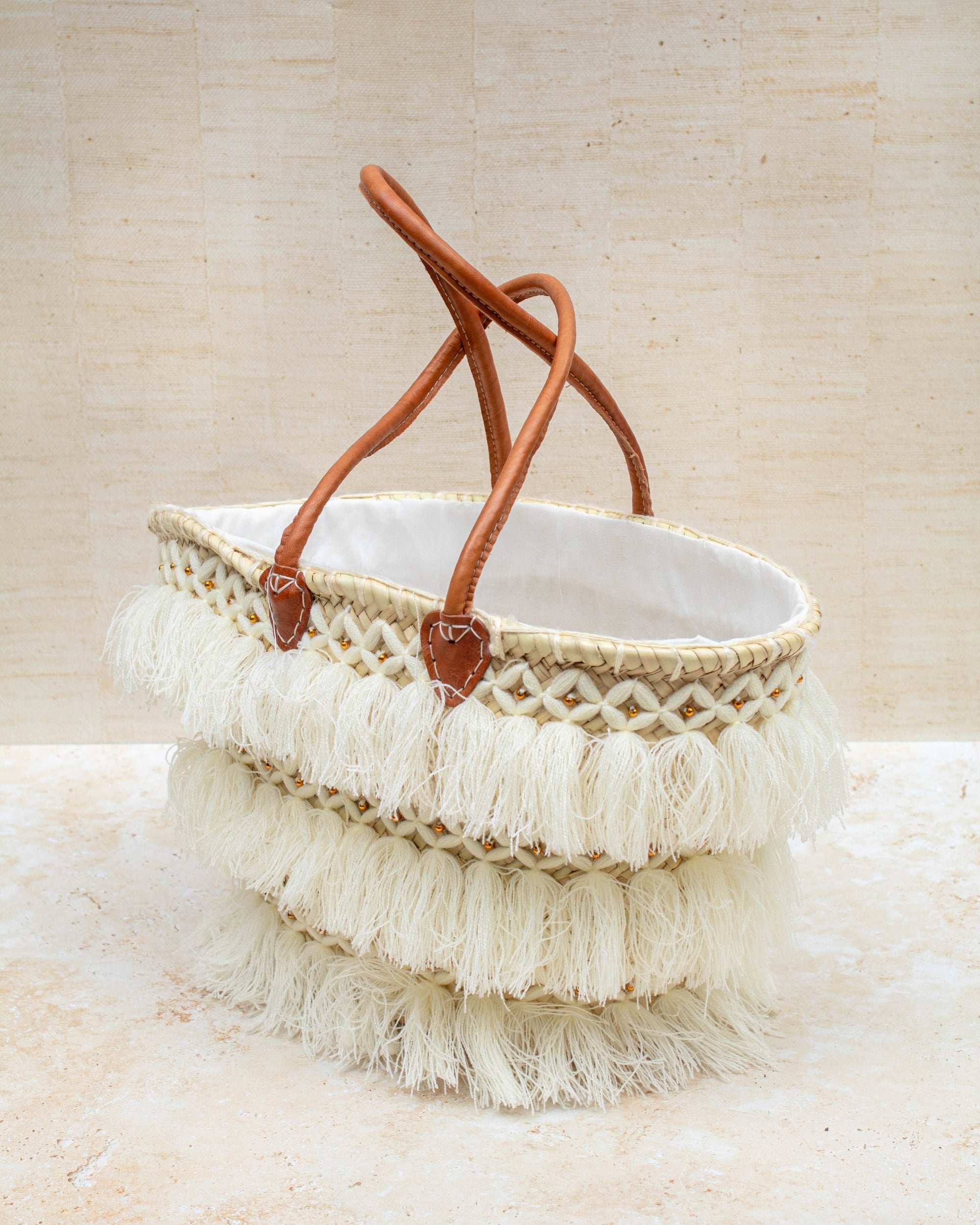 Beige woven bag with leather handles and white fringe on a beige background